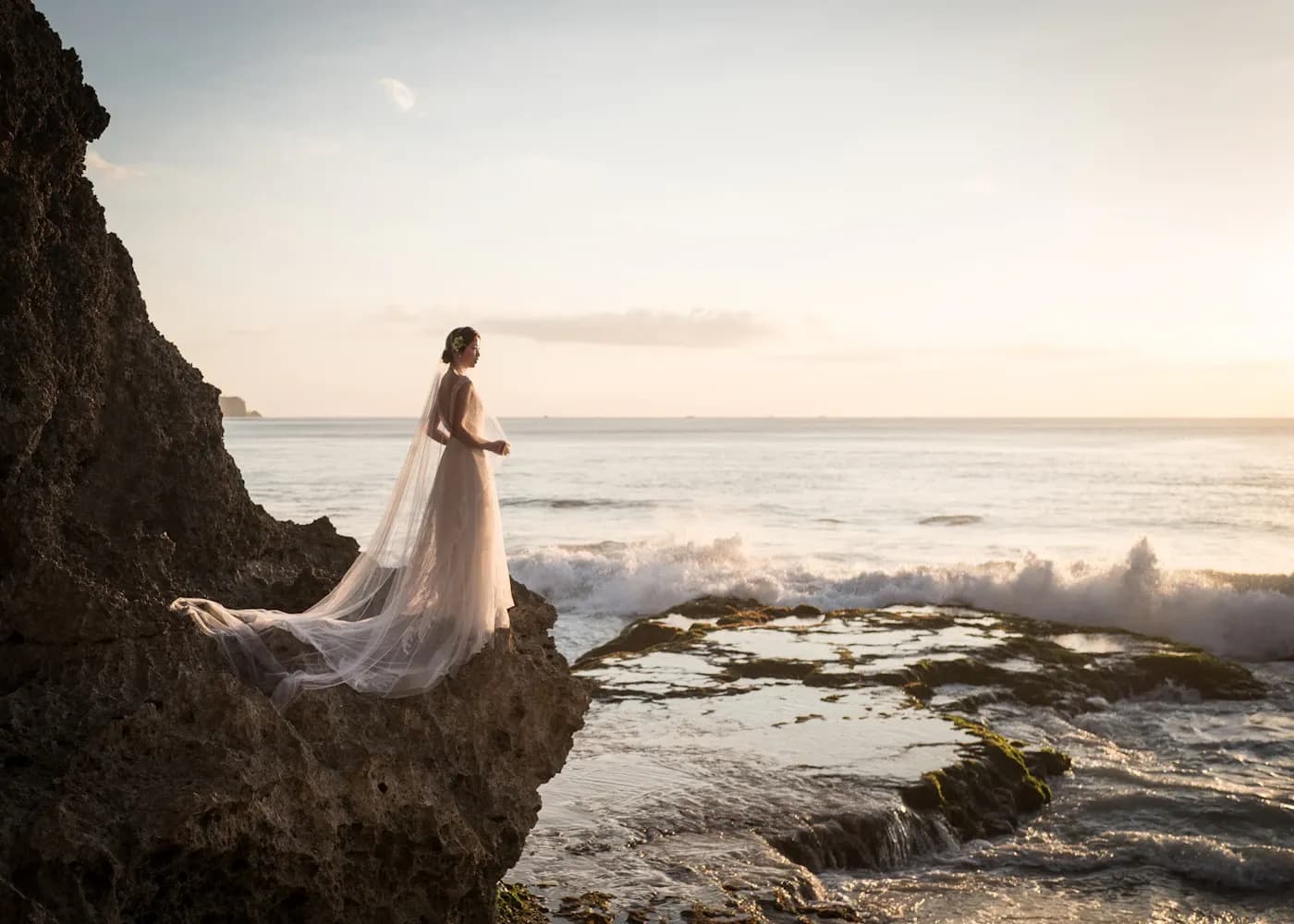 Bride in a gown standing on a rocky cliff by the sea at sunset